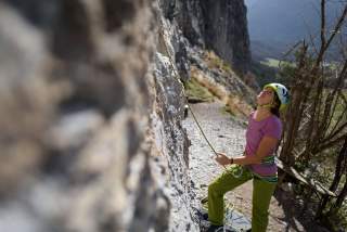 Eine Frau mit Kletterhelm und Kletterausrüstung sichert am Fuße einer Felswand einen Kletterer und blickt nach oben. Im Hintergrund sind Berge und ein felsiger Weg zu sehen.