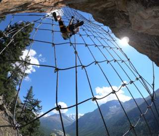 Eine Person klettert an einem Seilnetz an einem Felsen mit Bergpanorama und klarem Himmel im Hintergrund. Die Sonne scheint am rechten Bildrand hinter dem Felsen.