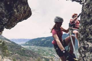 Zwei Personen klettern mit Kletterausrüstung an einer felsigen Steilwand, im Hintergrund ist ein Tal mit Bergen zu sehen.