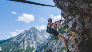 Ein Mann hängt stark am Felsen im Klettersteig; im Hintergrund zeigen steinerne Gipfel gen Himmel.