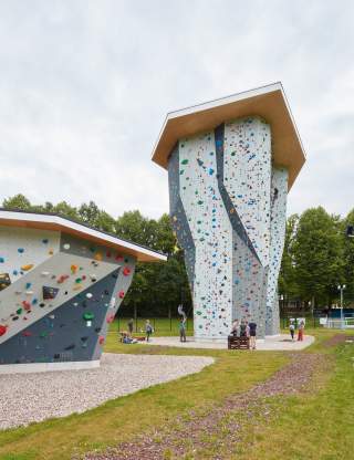 Zwei große, moderne Kletterwände im Freien mit bunten Griffen, umgeben von Wiese und Bäumen; mehrere Menschen klettern oder stehen am Boden.