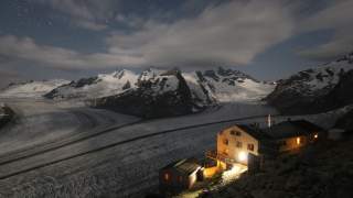 Nachtaufnahme einer Berghütte mit beleuchteten Fenstern vor einem Gletscher und schneebedeckten Bergen unter einem teilweise bewölkten, sternenklaren Himmel.