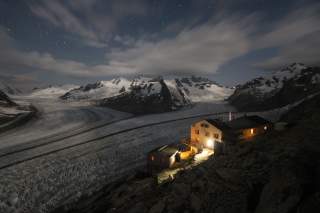 Berglandschaft bei Nacht mit schneebedeckten Gipfeln, eisigem Gletscher und einer beleuchteten Berghütte im Vordergrund. Ein klarer Sternenhimmel mit wenigen Wolken ist sichtbar.