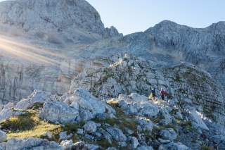 Zwei Wanderer gehen über felsiges Gelände in einer alpinen Berglandschaft bei Sonnenschein.