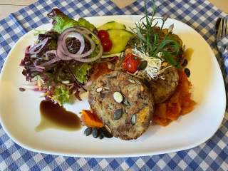 Weißer Teller mit zwei gebratenen Gemüsepuffern, frischem Salat mit roten Zwiebeln und Gurkenscheiben sowie einem Klecks dunkler Soße, garniert mit Kräutern und Sprossen.
