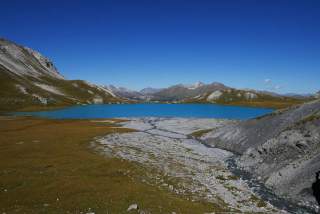 Bergsee mit blauem Wasser umgeben von grünen und felsigen Hügeln unter klarem, blauem Himmel. Im Vordergrund fließt ein kleiner Bach durch eine steinige Landschaft.