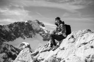 Schwarz-weiß-Foto einer lächelnden Person mit Rucksack, die auf einem felsigen Berghang sitzt, im Hintergrund schneebedeckte Berggipfel.