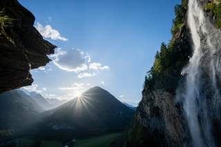 Sonnenaufgang hinter einem Berg mit Wasserfall und Felsen im Vordergrund, blauer Himmel mit wenigen Wolken.