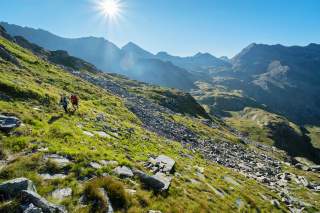 Zwei Wanderer gehen bei Sonnenschein einen steinigen, grünen Berghang entlang, im Hintergrund hohe Berge.