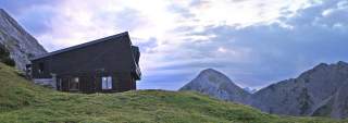 Holzhütte auf einer grünen Bergwiese mit imposanten Bergen und bewölktem Himmel im Hintergrund.