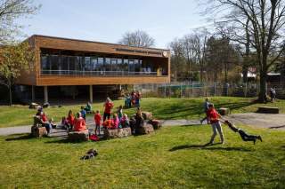 Kinder spielen und sitzen auf Steinen vor einem modernen Holzgebäude, umgeben von Rasenfläche und Bäumen.