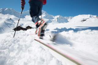 Eine Person fährt mit Skiern einen schneebedeckten Hang hinunter, im Hintergrund sind schneebedeckte Berge und Häuser unter klarem, blauem Himmel zu sehen.