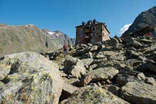 Zwei Wanderer steigen über felsiges Gelände auf eine Berghütte zu, umgeben von hohen Bergen unter blauem Himmel.