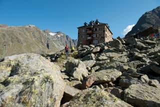 Zwei Wanderer gehen über einen steinigen Weg auf eine Berghütte inmitten einer felsigen Hochgebirgslandschaft zu.