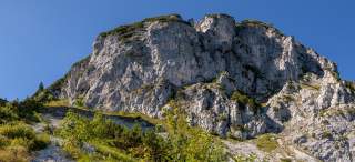 Felsiger Gebirgshang mit vereinzeltem Grünbewuchs unter klarem blauem Himmel. Vor dem Berg sind kleine Bäume und Sträucher auf einer steinigen Wiese zu sehen.
