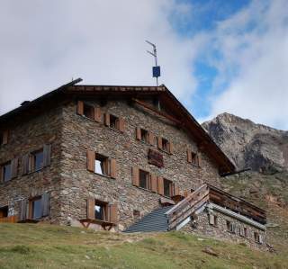 Steinhütte mit Holzfenstern auf einer Wiese vor einem Berghang, teils bewölkter Himmel.