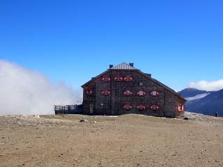 Berghütte mit roten Fensterläden steht auf kargem Gelände unter klarem blauem Himmel, im Hintergrund sind Berge und Wolken zu sehen.
