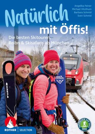 Zwei lächelnde Frauen in Skibekleidung und Mützen mit Skiern auf dem Rücken stehen vor einem roten Zug auf schneebedeckten Bahngleisen.