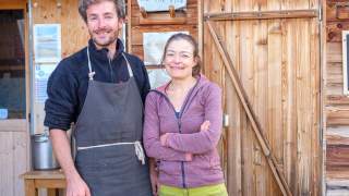 Zwei lächelnde Personen, ein Mann mit Schürze und eine Frau in Sportjacke, stehen vor einer Holzhütte mit einem Schild "LA VALETTE REFUGE P.N.V.".