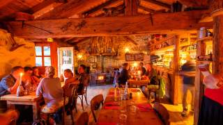 Gemütliches Restaurant mit Holzmöbeln und Kamin, in dem mehrere Gruppen von Gästen bei Kerzenlicht speisen. Die warme Beleuchtung und rustikale Einrichtung schaffen eine einladende Atmosphäre.