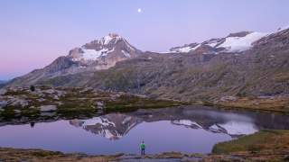 Eine Person steht an einem ruhigen Bergsee, in dem sich schneebedeckte Gipfel und ein blasser Mond im Dämmerlicht spiegeln.