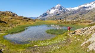 Wanderer mit rotem Rucksack geht an einem kleinen Bergsee entlang, im Hintergrund schneebedeckte Berge unter klarem blauem Himmel.