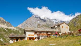 Berggasthof in den Alpen mit schneebedecktem Berg und Wolken im Hintergrund, umgeben von grüner Wiese und Wanderern.