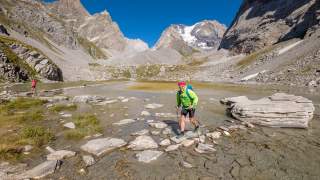 Wanderer mit grünem Rucksack und Trekkingstöcken überqueren auf einem felsigen Pfad einen flachen Gebirgsbach, umgeben von steilen Bergen und blauem Himmel.