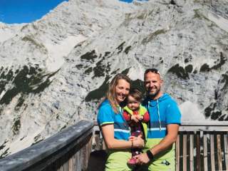 Eine Familie mit zwei Erwachsenen und einem Kleinkind steht lächelnd auf einer Aussichtsplattform vor einem felsigen Bergpanorama bei sonnigem Wetter.