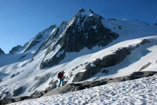 Eine Person mit Rucksack wandert auf verschneitem Berggelände vor einem steilen, schneebedeckten Berg unter blauem Himmel.