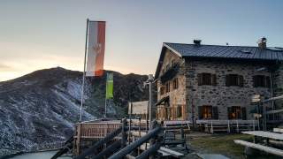 Steinernes Berghaus mit Holzgeländer und Sitzbänken vor schneebedecktem Berg im Hintergrund bei Sonnenuntergang, zwei Fahnen wehen im Wind.