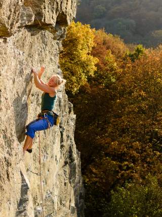 Eine Frau klettert an einer steilen Felswand, im Hintergrund sind herbstlich gefärbte Bäume zu sehen.