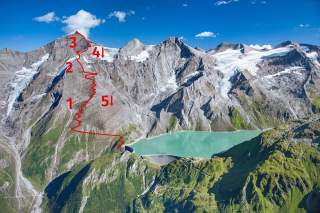 Luftaufnahme einer Berglandschaft mit einem Stausee, auf dem der Aufstiegsweg zum Gipfel in fünf Abschnitten rot markiert ist.