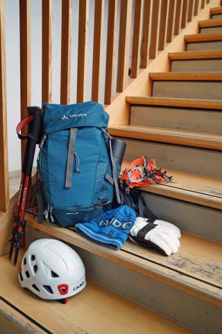 Rucksack, Bergsteigerausrüstung mit Helm, Handschuhen, Steigeisen, Mütze und Wanderstöcken liegen auf einer Holztreppe.