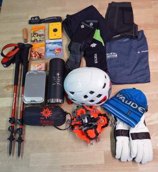 Ausrüstung für Berg- oder Wintertouren, darunter Helm, Steigeisen, Wanderstöcke, Handschuhe, Mütze, Thermoskanne, warme Kleidung, Lawinensicherheitsausrüstung und Snacks.