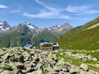 Berghütte mit Solarpanelen umgeben von Felsen und grasbewachsenen Hängen, im Hintergrund schneebedeckte Alpenberge unter blauem Himmel.