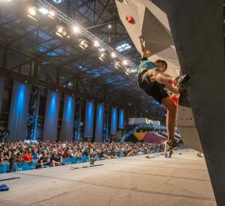Ein Boulderer klettert an einer Indoor-Kletterwand vor großem Publikum in einer Halle mit Bühnenbeleuchtung.