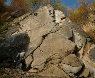 Mehrere Personen klettern an einer felsigen Kletterwand im Freien, während andere am Fuße des Felsens sichern, umgeben von herbstlichen Bäumen.