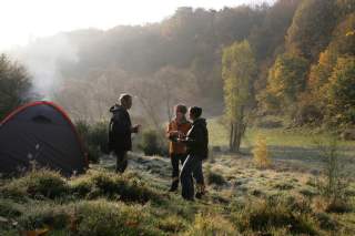 Drei Personen stehen auf einer Wiese neben einem Zelt und trinken aus Tassen, umgeben von herbstlichen Bäumen und leichtem Morgennebel.