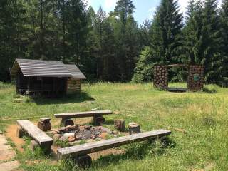 Eine Feuerstelle mit drei rustikalen Holzbänken steht auf einer Lichtung, links davon ein kleines Holzhäuschen und rechts davon ein Holzrahmen aus Baumstämmen, umgeben von hohen Nadelbäumen.