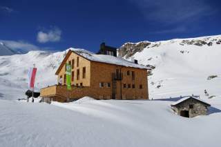 Hütte und kleine Steinhütte sind von Schnee bedeckt, im Hintergrund schneebedeckte Berge unter klarem blauem Himmel.