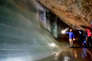 Eine Gruppe von Personen mit Helmen und Taschenlampen steht in einer Eishöhle, während eine Person vorne eine Lichtquelle auf eine große Eisschicht an der Höhlenwand richtet.
