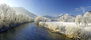Winterlandschaft mit einem zugefrorenen Fluss, schneebedeckten Bäumen und Bergen unter klarem blauem Himmel.