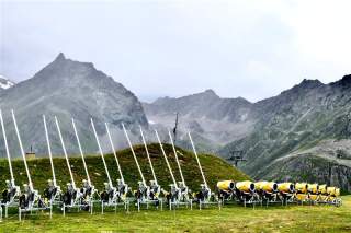 Reihe von Schneekanonen auf einer Wiese vor einer Bergkulisse.