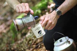 Person gießt Kaffee aus einer Espressokanne in eine Kaffeetasse mit Aufschrift „Waldschrat Tasse“ im Wald.