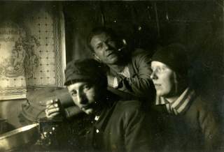Schwarzweißfoto von drei nachdenklich blickenden Personen in einer dunklen Umgebung, zwei Männer und eine Frau mit Kopfbedeckung. Im Hintergrund ist ein dekoratives Wandbild mit Engeln zu sehen.