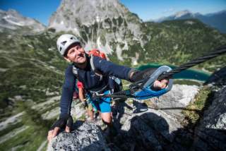 Ein Bergsteiger mit Helm und Kletterausrüstung hält sich an einem Sicherungsseil fest und steigt an einem Felsgrat in den Bergen empor, im Hintergrund sind weitere Kletterer und ein See zu sehen.