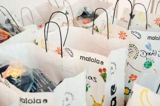 Mehrere weiße Papiertüten mit schwarzen Henkelgriffen, die mit bunten Zeichnungen und Schriftzügen bedruckt sind, gefüllt mit verpackten Gegenständen.