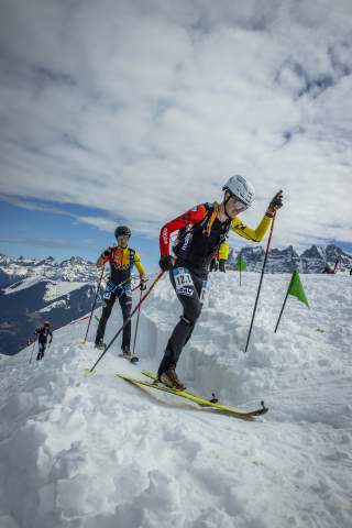 Mehrere Skibergsteiger in Rennkleidung mit Helmen und Skistöcken bewegen sich in einer verschneiten Berglandschaft entlang einer markierten Route.