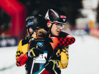 Zwei Skilangläufer in Helmen und Sportkleidung umarmen sich auf einer verschneiten Strecke.